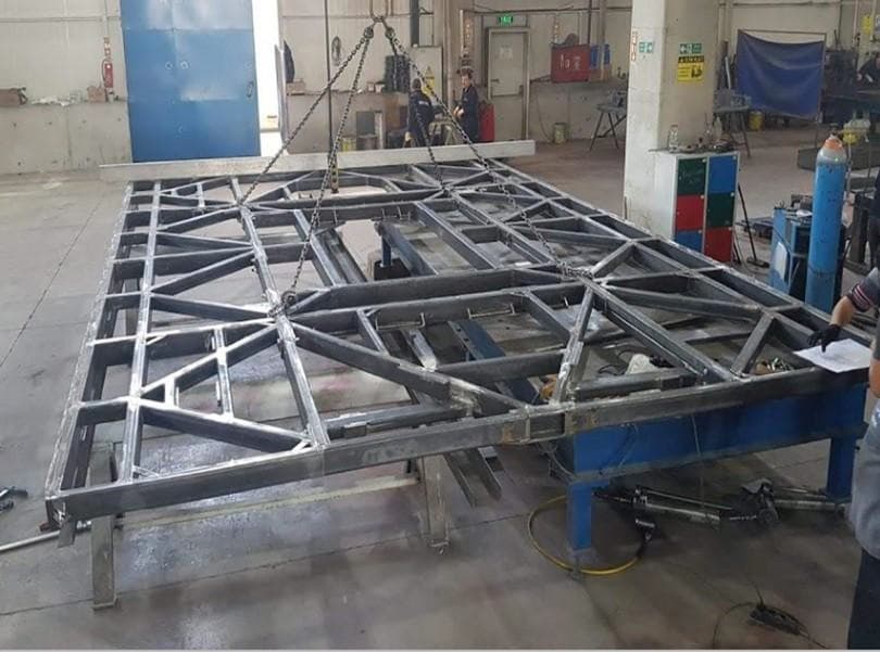 Welded steel platform frame being assembled and lifted by chain hoist in a fabrication workshop