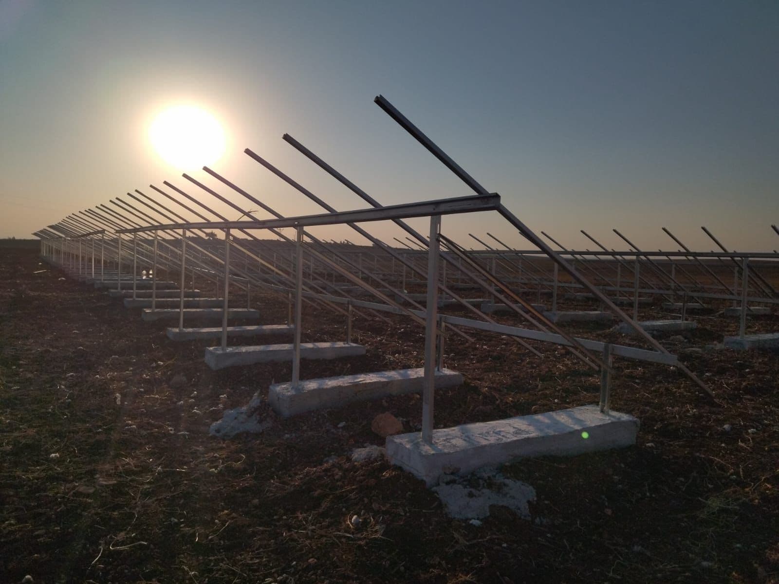 Rows of steel mounting frames for a ground-mounted solar panel array, backlit by the rising sun