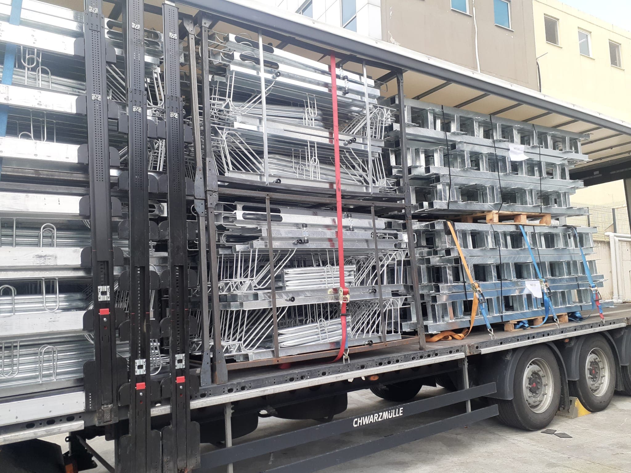 Flatbed truck loaded with galvanized steel rack frames and channel beams outside a facility